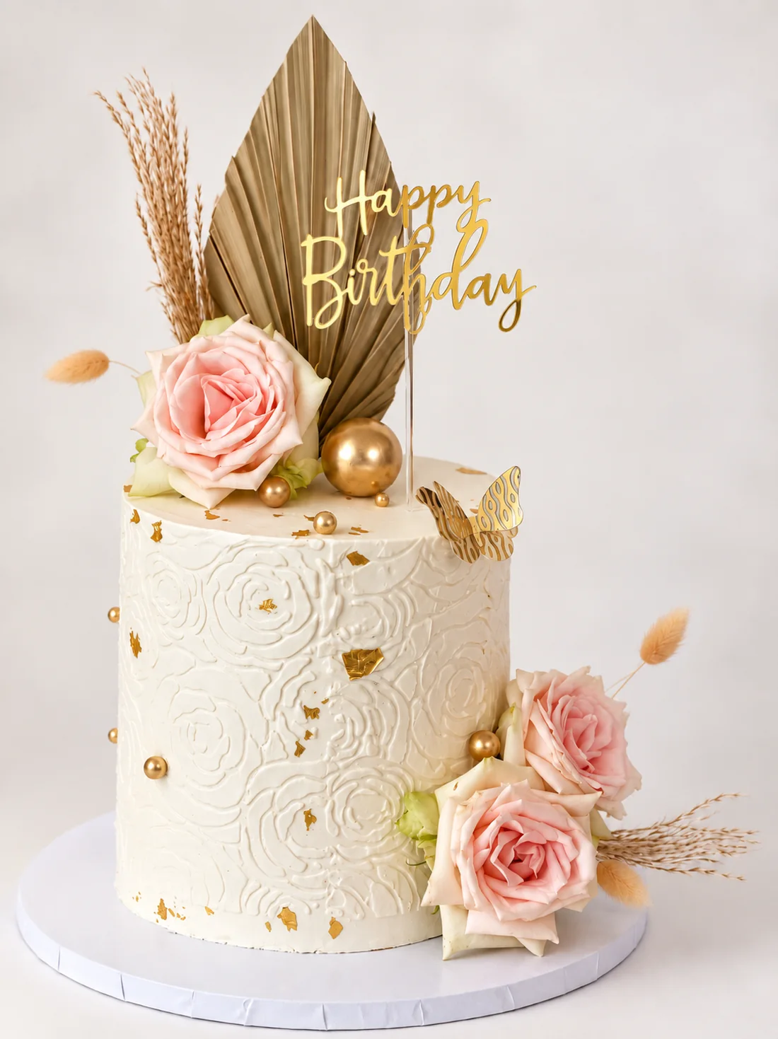 White textured cake with blush roses, dried palm leaf, pampas, gold spheres and a Happy Birthday topper